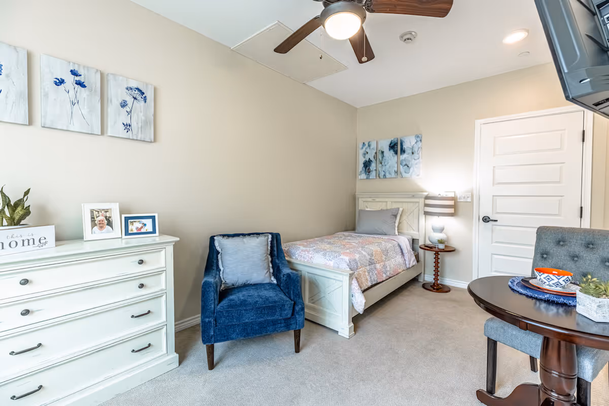 A cozy bedroom with a single bed covered in a pastel patchwork quilt, a blue upholstered armchair with a cushion, a white dresser with framed photos and a plant, a round wooden table with a bowl and a small plant, and a ceiling fan with a light. The walls are beige with blue floral artwork above the bed and dresser.