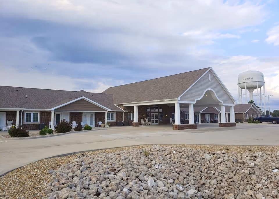 Exterior view of a single-story senior living facility building with a large covered entrance supported by white columns. The building has a brick facade with white trim and several windows and doors. There are rocking chairs placed near the entrance and along the building. In the foreground, there is a circular area filled with rocks, and in the background, a water tower labeled 'CHATHAM' is visible under a partly cloudy sky.