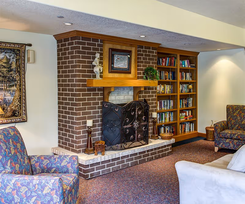 Cozy living room area with a brick fireplace featuring a decorative black metal screen. The fireplace mantel holds a small statue, a framed picture, and a potted plant. To the right of the fireplace is a wooden bookshelf filled with books. There are patterned armchairs and a white sofa arranged around the fireplace on a carpeted floor. A tapestry hangs on the wall to the left.