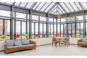 A bright and spacious indoor seating area with large floor-to-ceiling windows and a glass ceiling allowing natural light to fill the space. The room features two cushioned wooden sofas with patterned pillows and a small round table with four chairs. Potted plants with red and yellow flowers are placed along the window ledge.