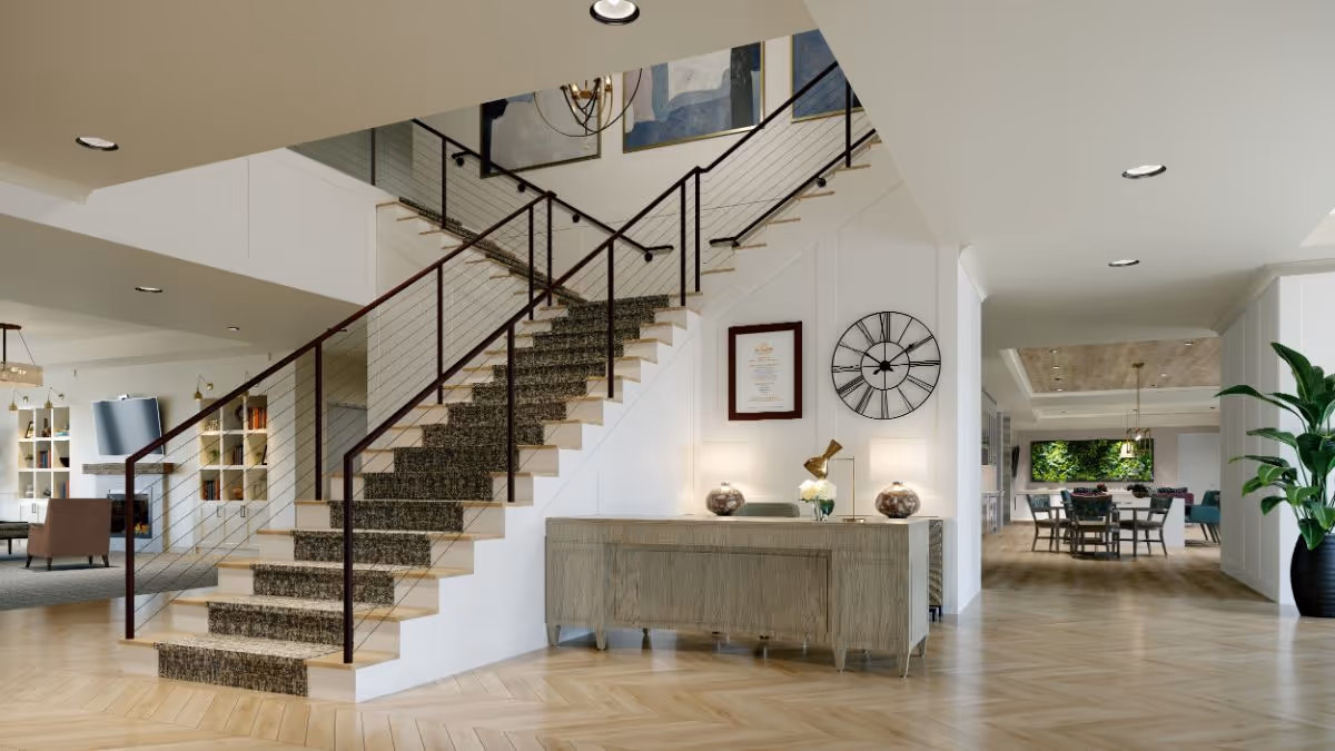 Interior view of a senior living facility showing a modern staircase with carpeted steps and metal railing. To the right of the staircase is a wooden console table with decorative items and a wall clock above it. In the background, there is a dining area with a table and chairs, and a living area with shelves and seating. The space features light wood flooring and recessed ceiling lights.