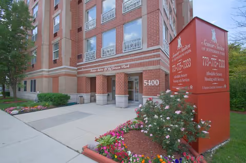 Exterior view of a multi-story brick senior living facility named Senior Suites of Jefferson Park with a red sign displaying the facility's name, phone number, and services. The entrance is surrounded by well-maintained flower beds and greenery.