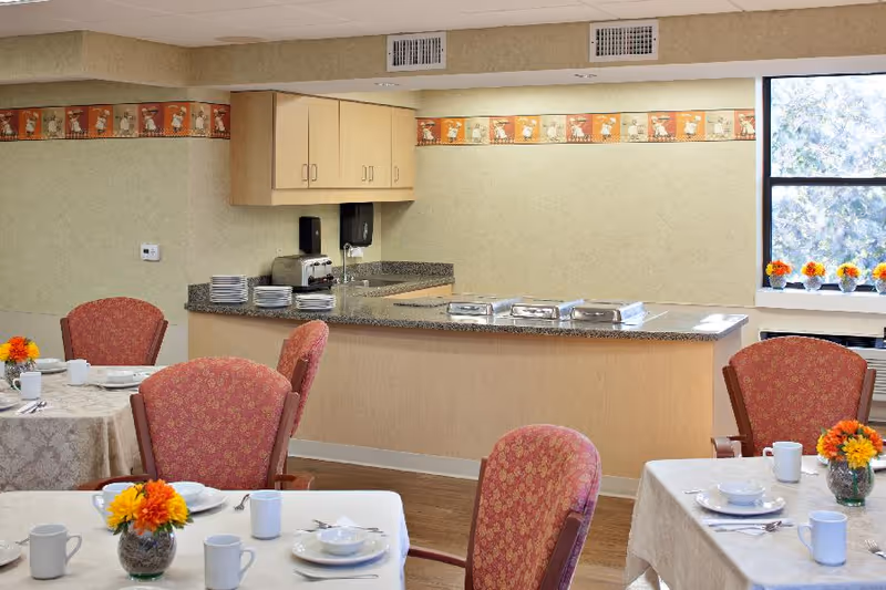 Dining room with table settings, floral centerpieces, upholstered chairs, and a serving counter with plates and chafing dishes.