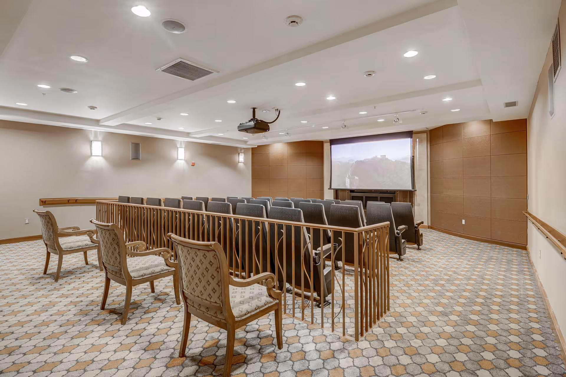 Carpeted screening room with rows of theater seats facing a projection screen and a few upholstered chairs in the foreground.