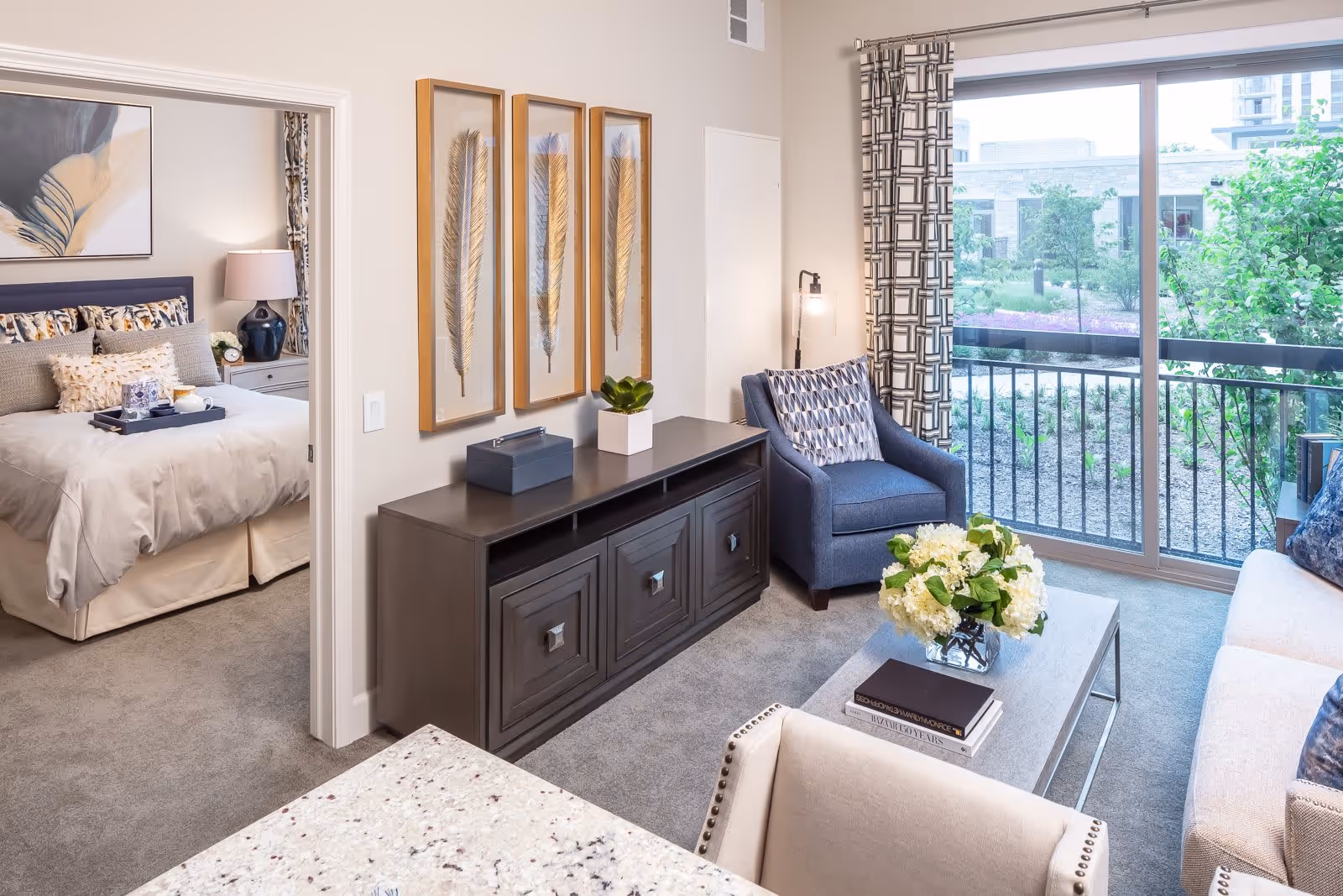 A bright and modern living room with a large window and sliding glass door overlooking a garden. The room features a beige sofa, a blue armchair with a patterned pillow, a coffee table with a vase of white flowers and books, and a dark wooden cabinet with decorative items. Adjacent to the living room is a bedroom with a neatly made bed, bedside table with a lamp, and wall art.
