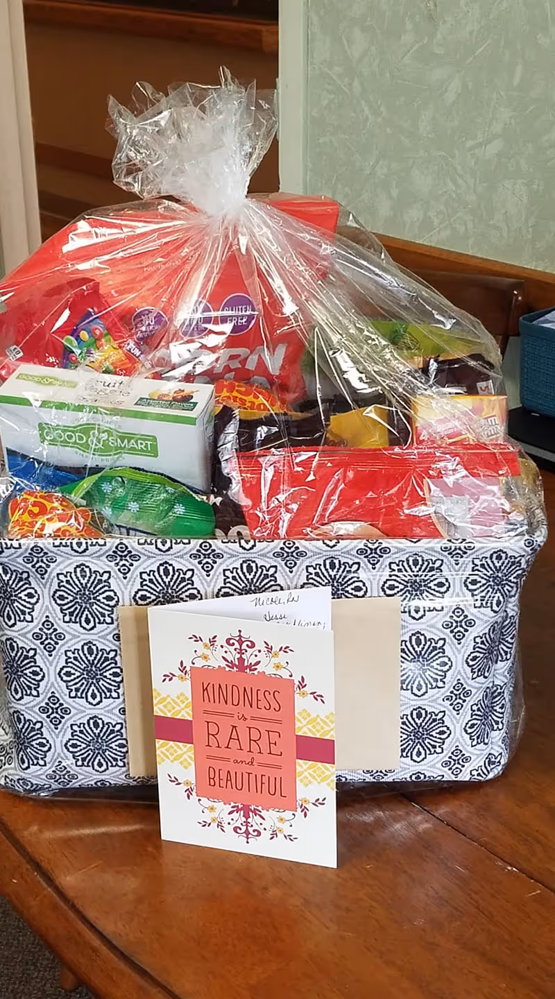 A gift basket wrapped in clear plastic containing various snacks and food items, placed on a wooden table. In front of the basket is a card with the message 'Kindness is rare and beautiful.' The basket has a black and white patterned fabric exterior.
