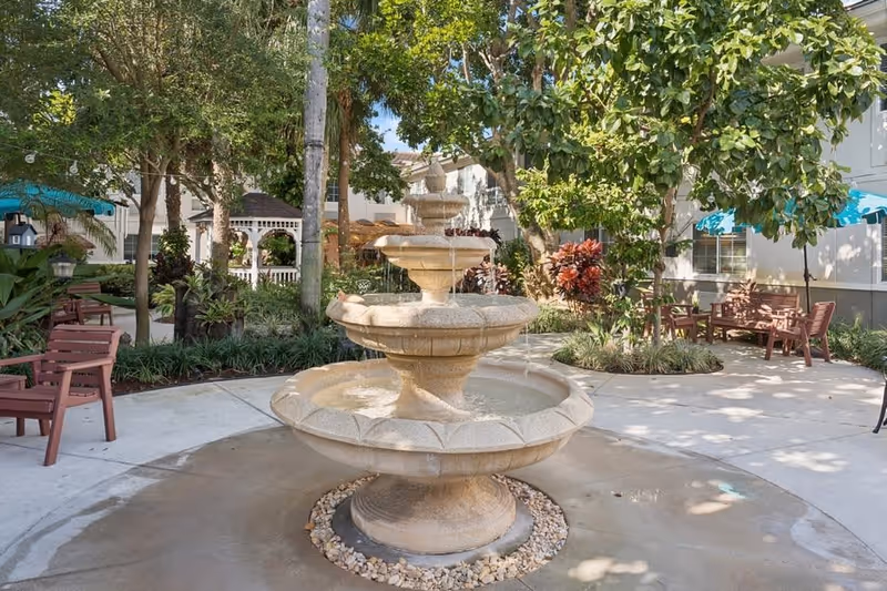 Tiered stone fountain centered in a shaded courtyard with benches, trees, and landscaping.