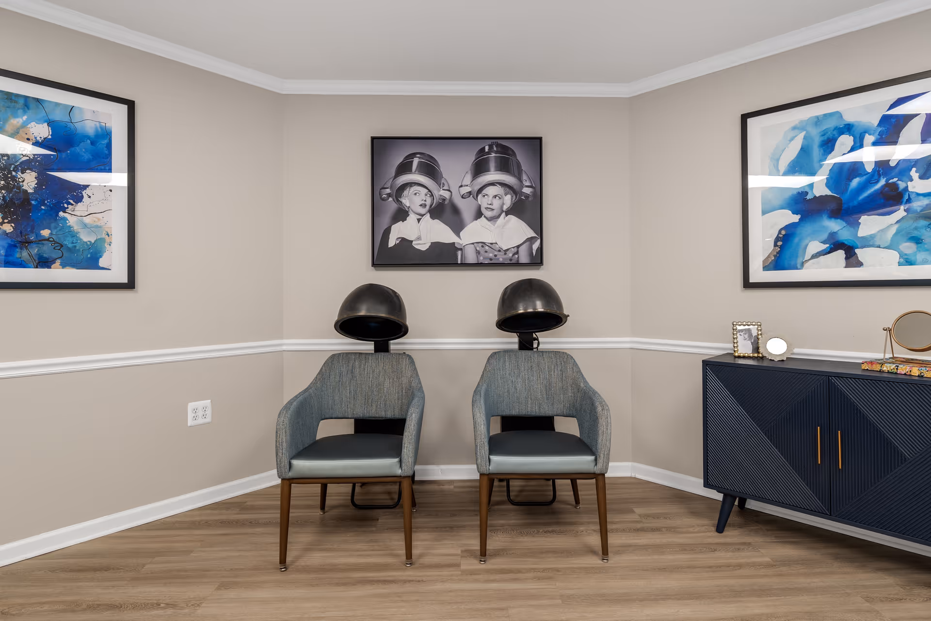 A small salon area with two vintage hair drying chairs with black hair dryers attached. The walls are beige with white trim, and there are three framed pictures on the walls: two abstract blue artworks on the left and right, and a black and white photo of two children under hair dryers in the center. A dark blue cabinet with gold handles is on the right side, holding a small mirror and a framed photo.