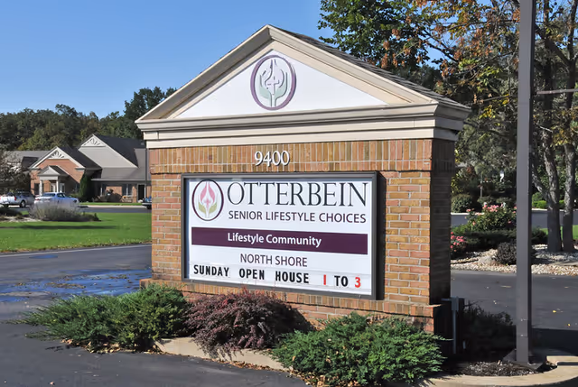 A brick sign at the entrance of Otterbein Marblehead SeniorLife Community with the address number 9400. The sign displays the community name and an announcement for a Sunday open house from 1 to 3. The sign is surrounded by landscaping with bushes and trees, and a parking lot is visible in the background.