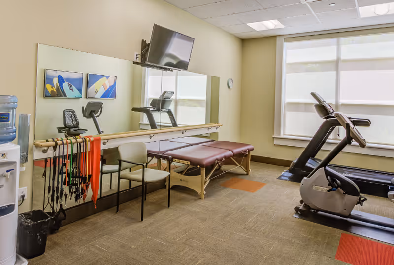 Well-lit fitness and therapy room with a treadmill and exercise bike, a padded therapy table, mirror, resistance bands and a wall-mounted TV.