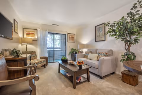 Cozy living room with a sofa, armchairs, coffee table, framed artwork, plants, and a sliding glass door.