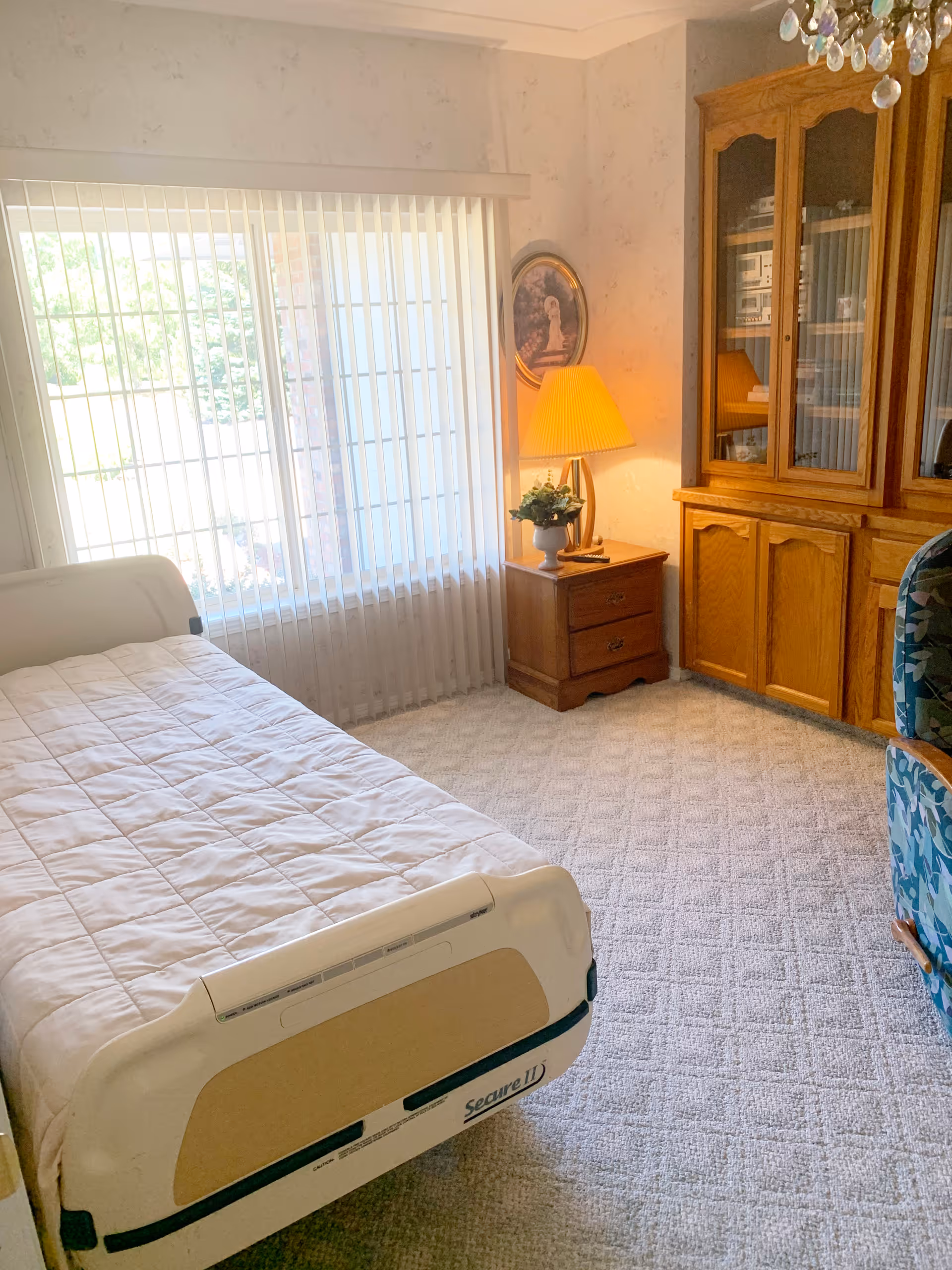 Sunny bedroom with a single adjustable bed, nightstand and lamp by a large window with vertical blinds, and a wooden cabinet and chair.