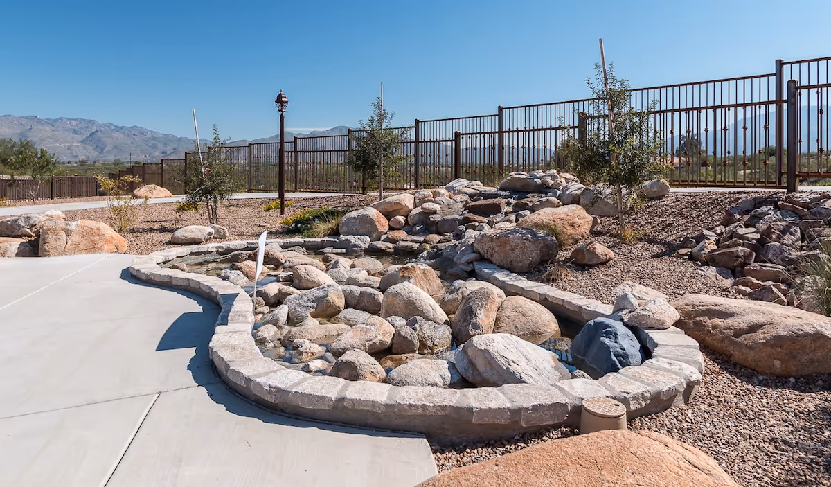 Landscaped outdoor area featuring a stone-lined water feature and dry creek beside a concrete walkway, fenced with mountains in the distance.