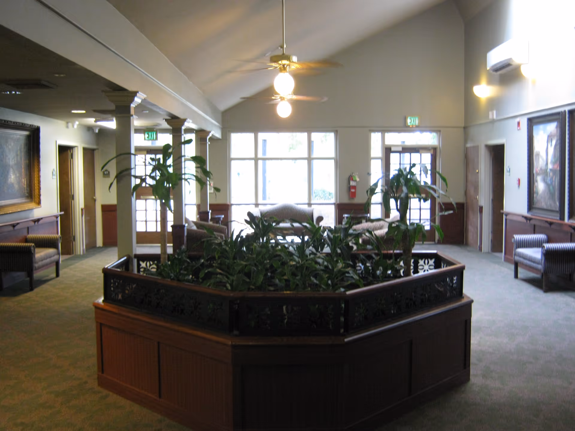 Interior lobby area with a central wooden planter filled with plants, seating, columns, and large windows in the background.