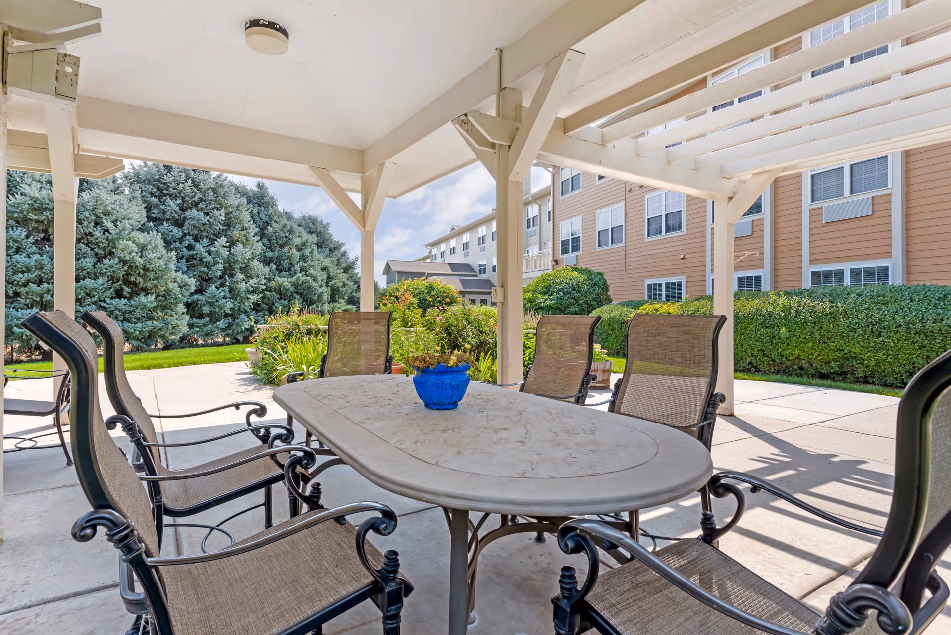Outdoor covered patio area with a large rectangular table surrounded by six metal and mesh chairs. A small blue planter with flowers is centered on the table. The patio is adjacent to a building with multiple windows and surrounded by green bushes and tall evergreen trees under a partly cloudy sky.