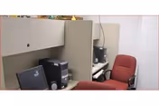 Small office cubicle area with a computer monitor, desktop computer, and a red office chair. The cubicle walls are beige and there is a second cubicle visible in the background with another computer setup.