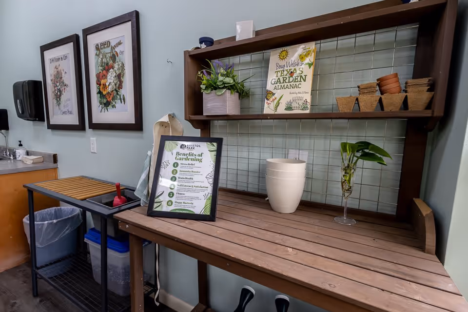 A gardening station inside a room with a wooden table and a shelf holding gardening pots, a plant, and a book titled 'Doug Welsh's Texas Garden Almanac'. A framed sign on the table lists the benefits of gardening. The wall has two framed floral prints, and there is a sink and a trash bin nearby.