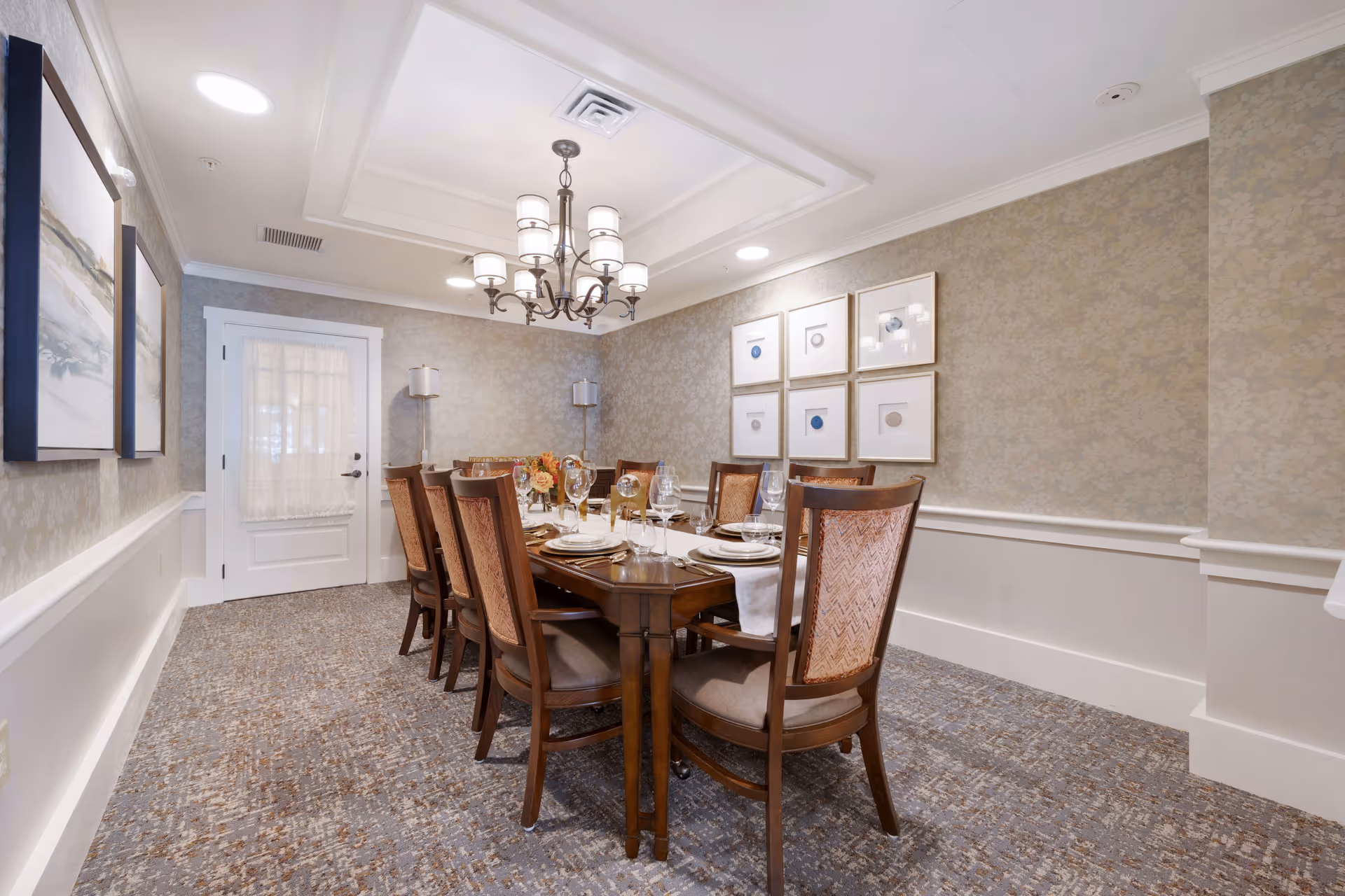 A formal dining room with a long wooden table set for eight people. The table is arranged with plates, wine glasses, and a centerpiece of flowers. The room has patterned wallpaper, carpeted floor, and a chandelier hanging from the ceiling. There are framed artworks on the walls and two floor lamps in the corner near a white door with a sheer curtain.