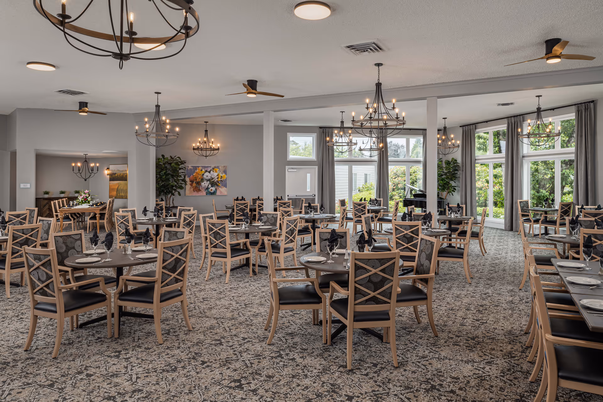 A spacious dining room with multiple round tables and wooden chairs with cushioned seats. Each table is set with plates, glasses, and black folded napkins. The room features large windows with gray curtains allowing natural light to fill the space. Several elegant chandeliers hang from the ceiling, and ceiling fans are also visible. There is a piano near the windows and some decorative plants and artwork on the walls.