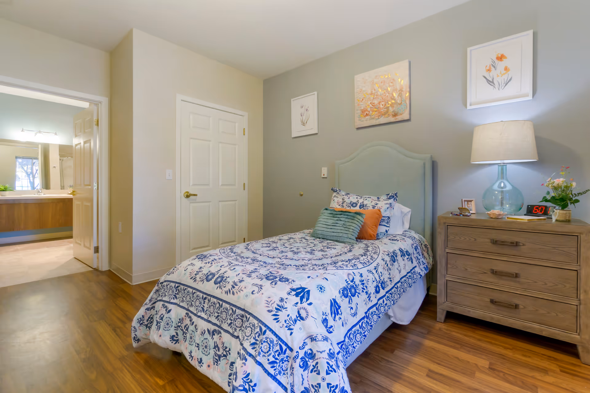 A cozy bedroom with a single bed featuring a blue and white floral patterned bedspread and multiple pillows. Next to the bed is a wooden nightstand with a blue glass lamp, a digital alarm clock showing 6:01, a small framed photo, and a vase with flowers. The walls are decorated with three framed floral artworks. A door leads to an adjacent bathroom with a large mirror and sink visible.