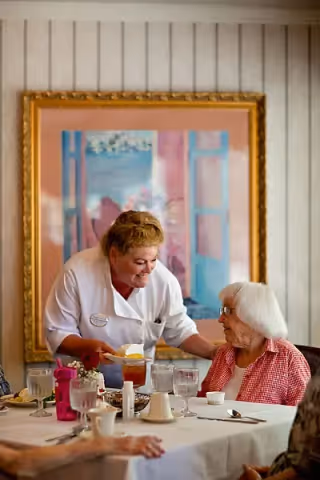 A caregiver in a white uniform serving food to an elderly woman with white hair and glasses seated at a dining table. The table is set with glasses, cups, and plates. A large framed painting with blue and pink tones hangs on the wall behind them.