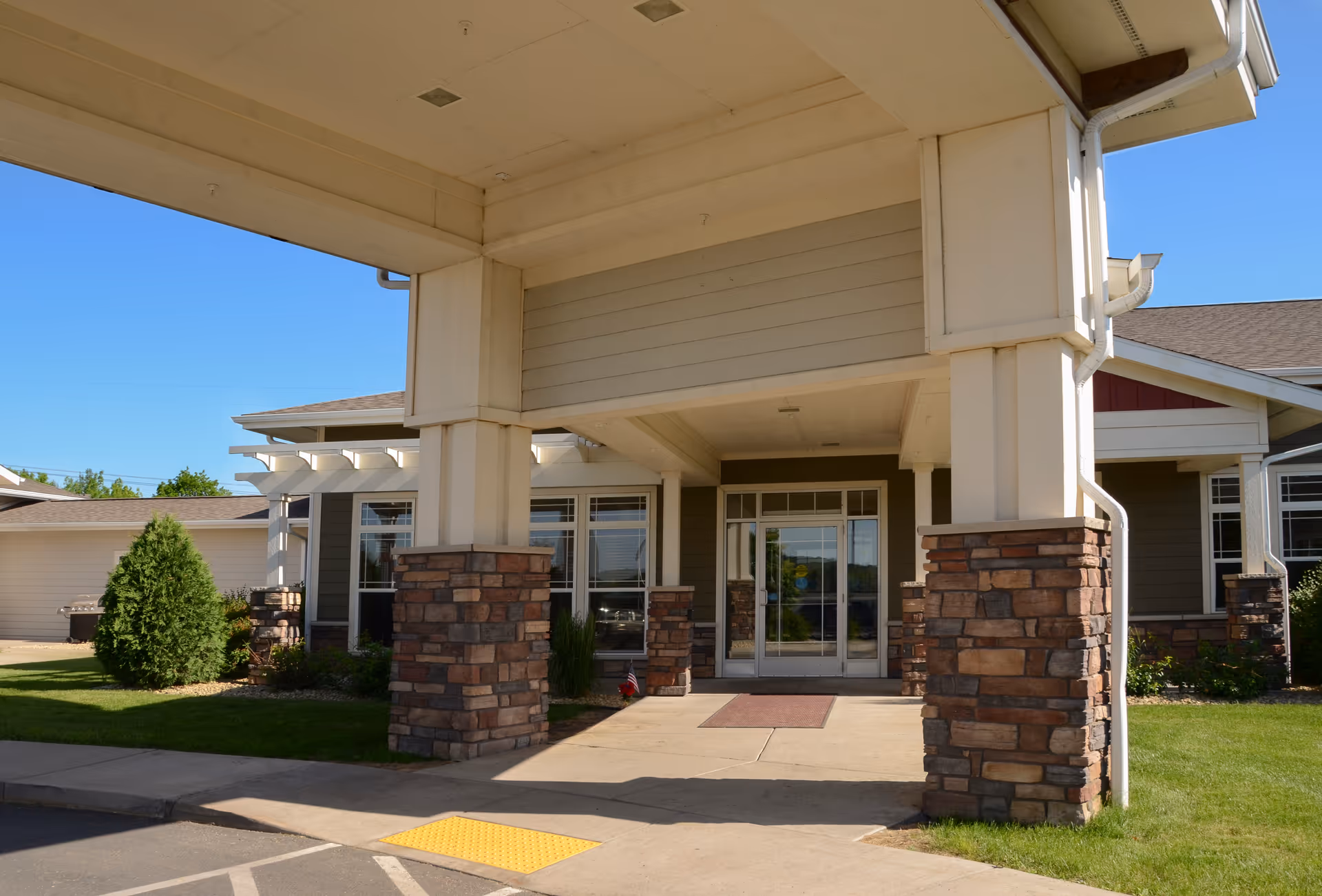 Entrance of a senior living facility with a covered drop-off area supported by stone pillars, glass double doors, and well-maintained landscaping including grass and shrubs under a clear blue sky.