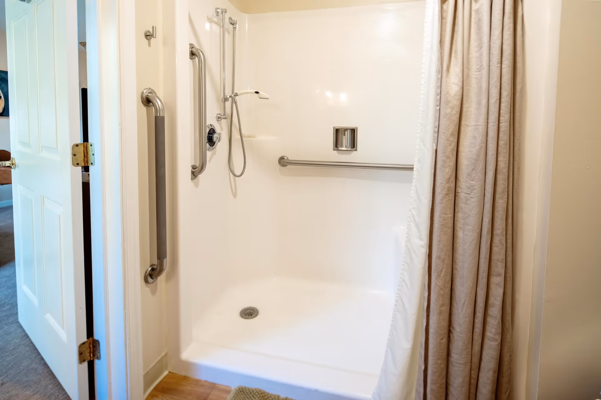 A clean, white walk-in shower with a handheld showerhead and multiple stainless steel grab bars for safety. The shower has a beige curtain partially drawn to the side. The door to the bathroom is open, showing part of an adjacent room with carpeted flooring.