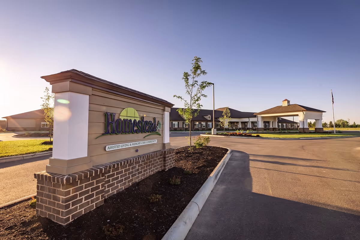 A 'Homestead' entrance sign and landscaped brick planter in front of a single-story assisted living building with a circular driveway under a clear sky.