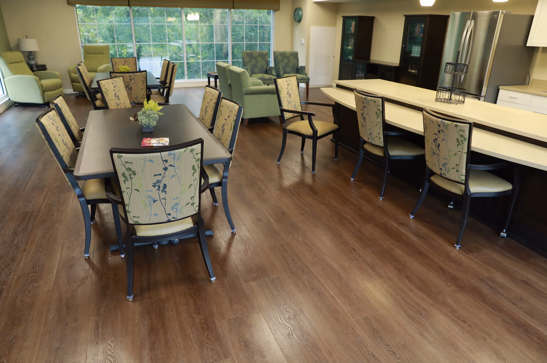 A spacious senior living common area with a dining table surrounded by chairs, a kitchen counter with additional chairs, and a seating area with armchairs near large windows showing greenery outside. The room has wood flooring and a bright, welcoming atmosphere.