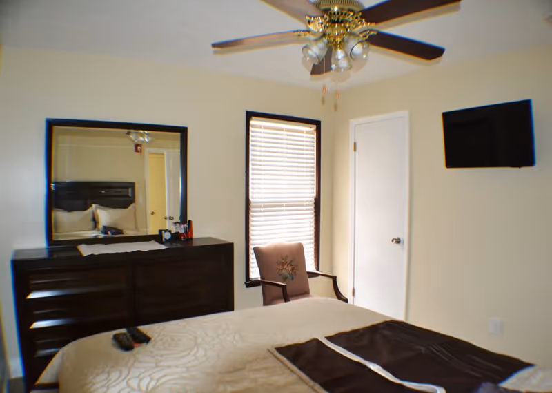 A small bedroom with a bed in the foreground, dresser and mirror, an armchair by a window, ceiling fan, and a wall-mounted TV.