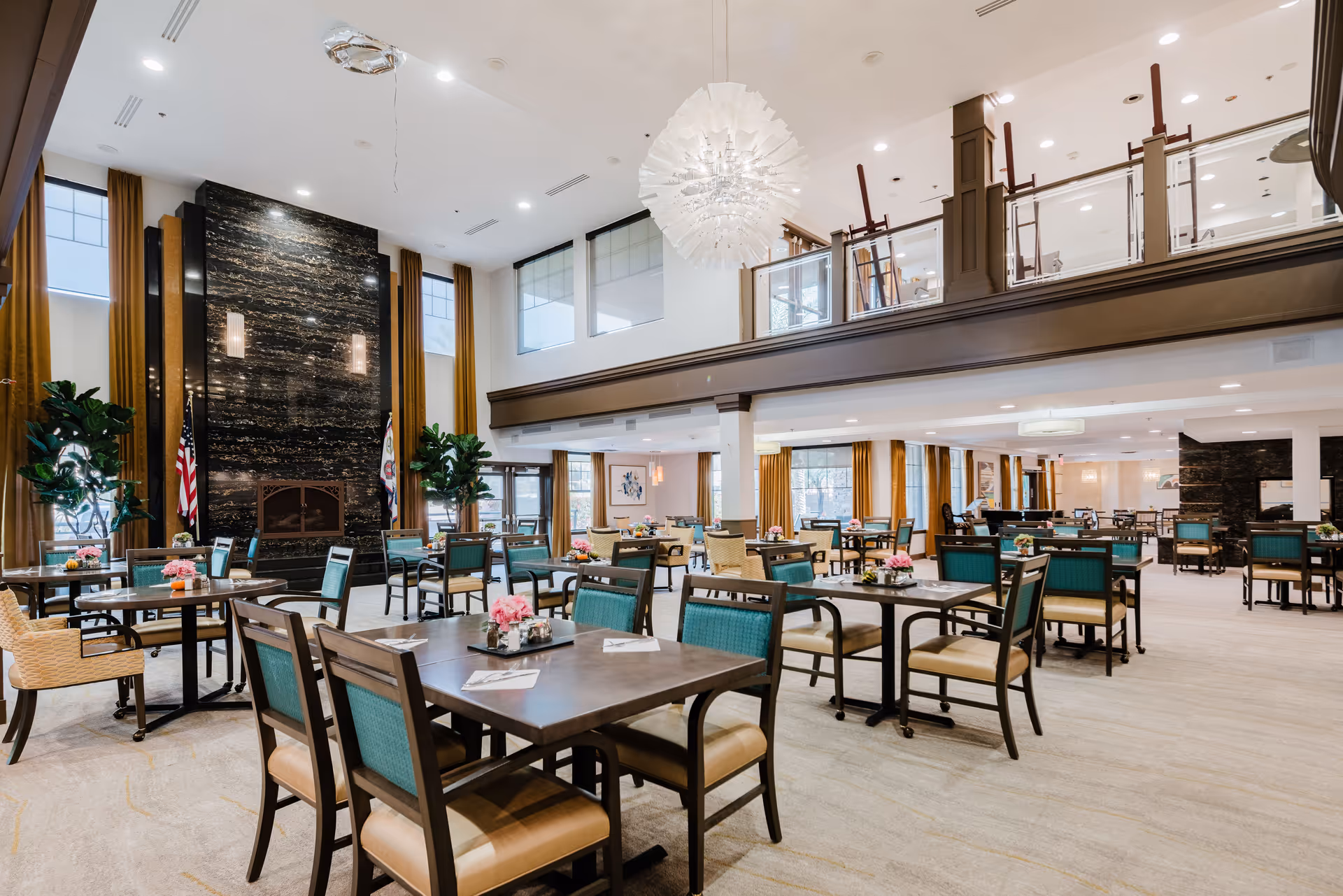 Spacious and well-lit dining room with multiple tables and chairs arranged neatly. The room features a high ceiling with a large modern chandelier, tall windows with gold curtains, a dark marble fireplace, and indoor plants. There is a mezzanine level with glass railings overlooking the dining area.