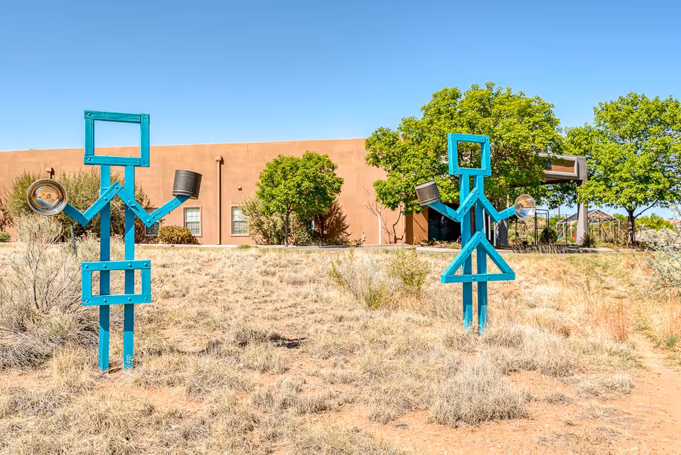 Two blue metal sculptures shaped like stylized human figures holding pots and pans, standing on dry grass with a brown building and green trees in the background under a clear blue sky.