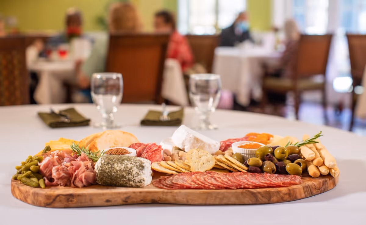 A wooden serving board with an assortment of cheeses, cured meats, crackers, olives, pickles, and breadsticks on a white tablecloth in a dining room setting with blurred people and tables in the background.