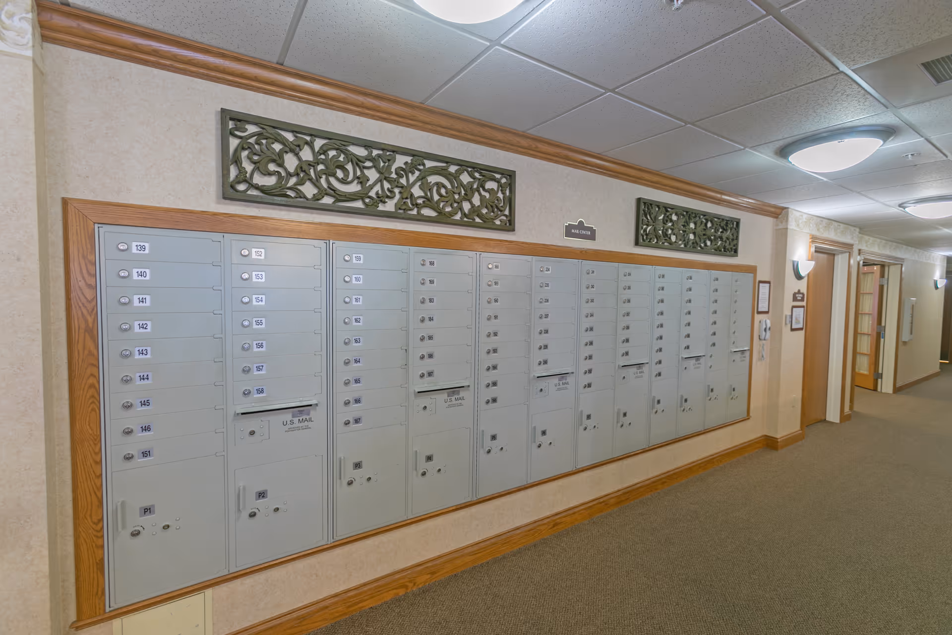 A hallway in a senior living facility with a row of mailboxes mounted on the wall. The mailboxes are numbered and have locks. The hallway has beige walls with wooden trim, decorative wall panels above the mailboxes, carpeted floor, and ceiling lights. There are doors and wall sconces along the hallway.
