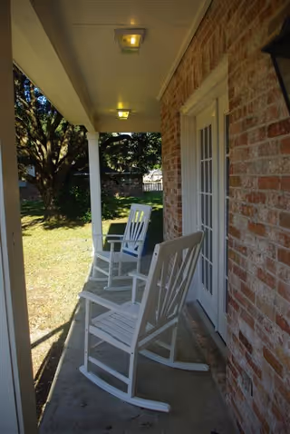 A covered brick porch with two white rocking chairs facing a grassy yard and trees.