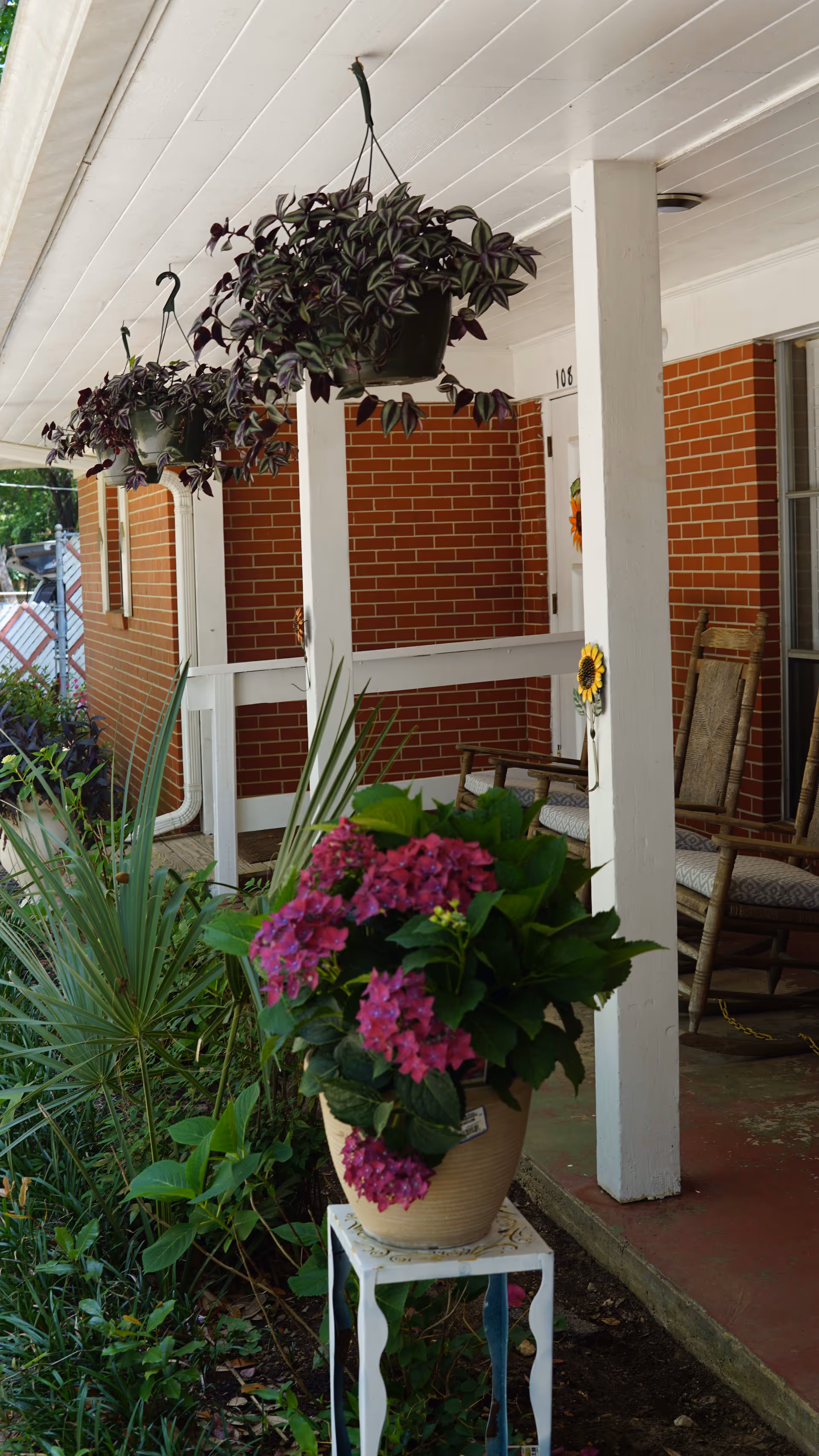 A porch area with white wooden pillars and a white ceiling, featuring hanging potted plants with green and purple leaves. There is a large potted plant with vibrant pink flowers placed on a small white stand in the foreground. The porch has a red brick wall with a door numbered 108, decorated with a sunflower ornament. Two wooden rocking chairs with cushions are positioned on the porch floor.