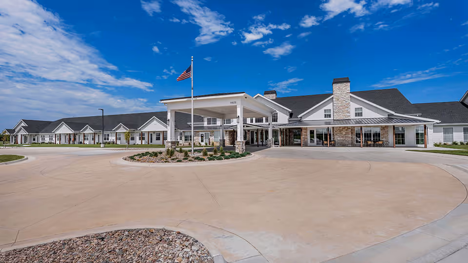 Exterior front view of a senior living building with a covered entrance, circular driveway, landscaped island, and an American flag under a blue sky.