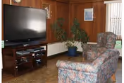 A cozy living room area with wood-paneled walls, a large flat-screen TV on a wooden stand, a potted plant, a floral-patterned armchair, and a matching ottoman. There is a window with vertical blinds allowing natural light into the room.