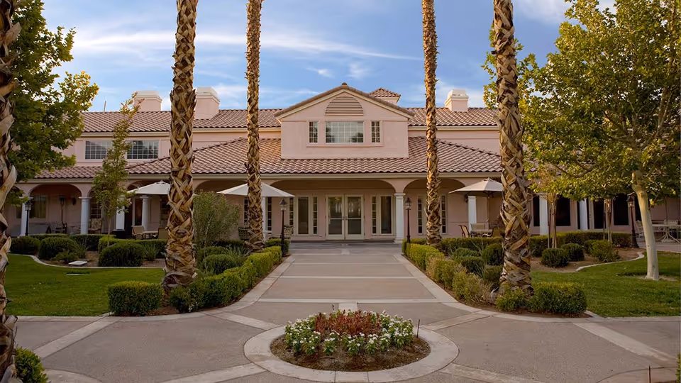 Front exterior view of a senior living facility with a tiled roof, multiple windows, and a central entrance. The building is surrounded by palm trees, manicured bushes, green lawns, and outdoor seating areas with umbrellas under a partly cloudy sky.