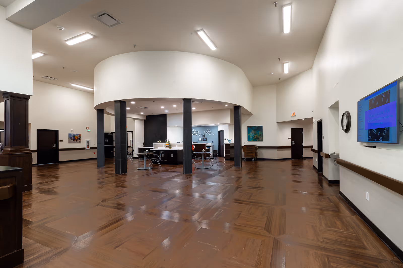 A spacious interior area with polished wooden floors and high ceilings. The room features a circular central section supported by black pillars, with tables and chairs arranged inside. Walls are painted white with some artwork and a wall-mounted TV visible. Several doors and a clock are also present along the walls.