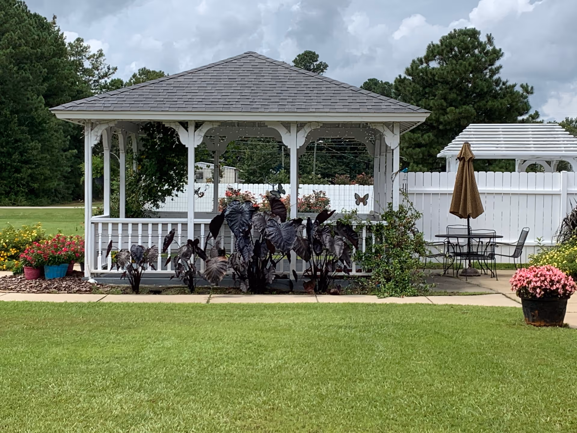A white gazebo with a shingled roof is situated in a well-maintained garden area with green grass and various flowering plants. To the right of the gazebo, there is a patio area with a table, chairs, and a closed brown umbrella. A white fence and trees are visible in the background under a cloudy sky.