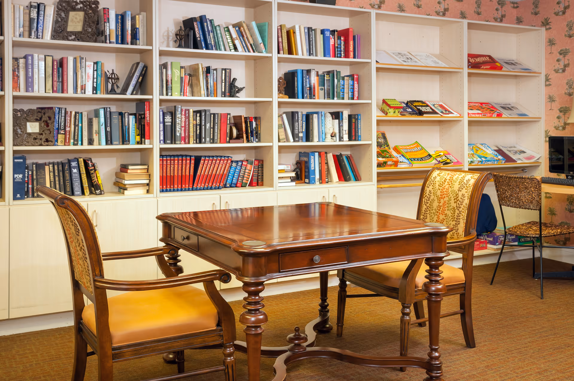 A cozy reading or activity room with a wooden table and two upholstered chairs in the foreground. Behind the table is a large white bookshelf filled with books, magazines, and board games. The room has warm lighting and patterned wallpaper on one wall.