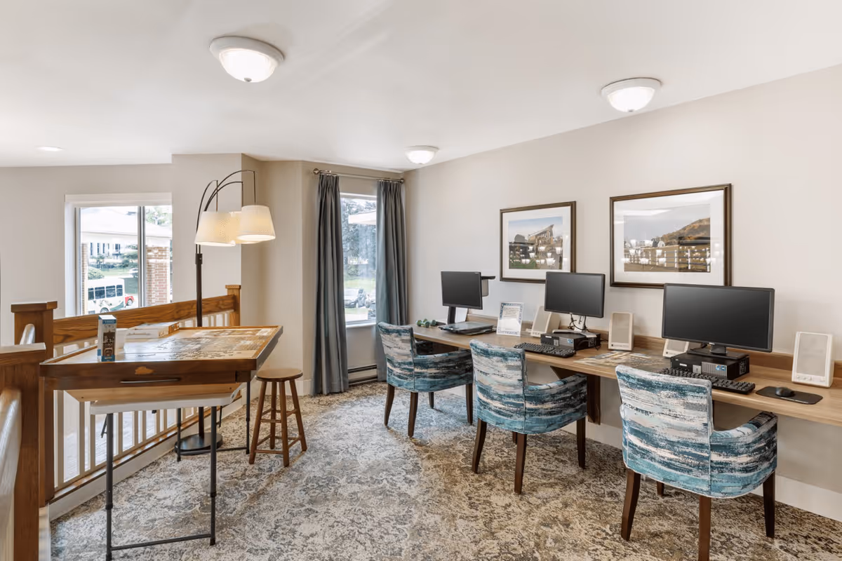 A bright room with three computer workstations along a wooden desk, each with a monitor, keyboard, and mouse. Three blue patterned chairs are positioned in front of the workstations. To the left, there is a wooden drafting table with a stool and a floor lamp with two lampshades. Two framed landscape pictures hang on the wall above the desk. A window with gray curtains lets in natural light.