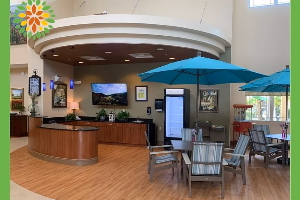 Interior view of a common area in Seasons Belleair Memory Care featuring a curved wooden reception desk, a wall-mounted TV, framed artwork, and a seating area with tables, chairs, and blue umbrellas. The space has wood flooring and decorative plants.