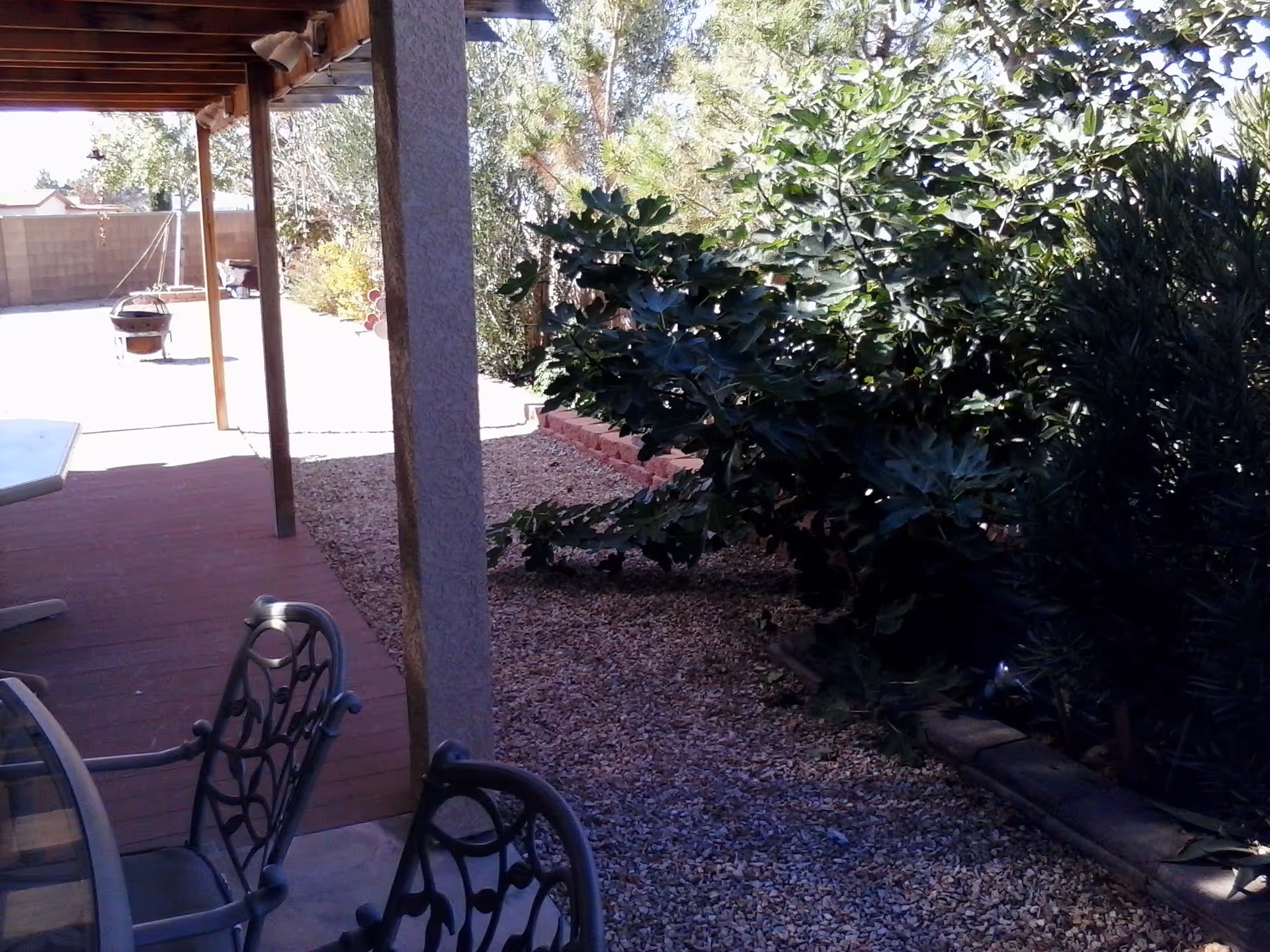 Outdoor patio area with a covered wooden pergola, metal chairs with decorative backs, a glass-top table, gravel ground, and lush green bushes along the side. A fire pit and a brick wall are visible in the background.