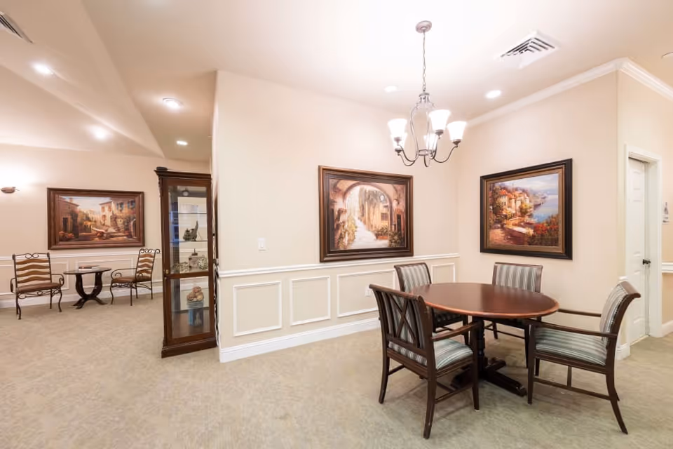 Small communal dining area with a round table and four chairs, framed artwork, a chandelier, and a display cabinet.