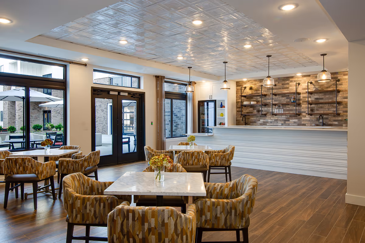 Bright communal dining area with marble-topped tables, patterned chairs, a long bar counter with pendant lights, and doors opening to an outdoor patio.
