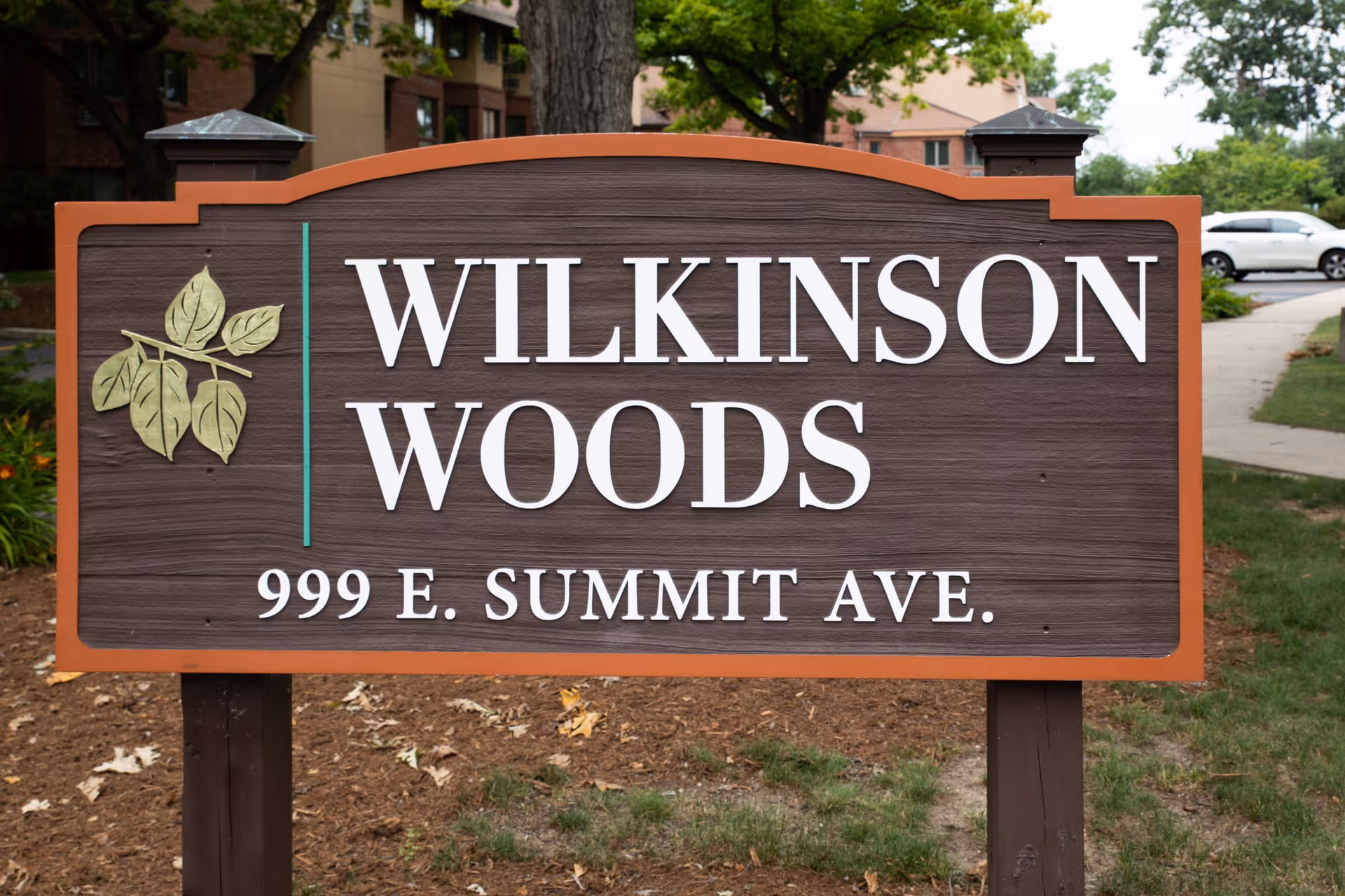 A wooden outdoor sign that reads "WILKINSON WOODS 999 E. SUMMIT AVE." with a leaf logo in front of the community.
