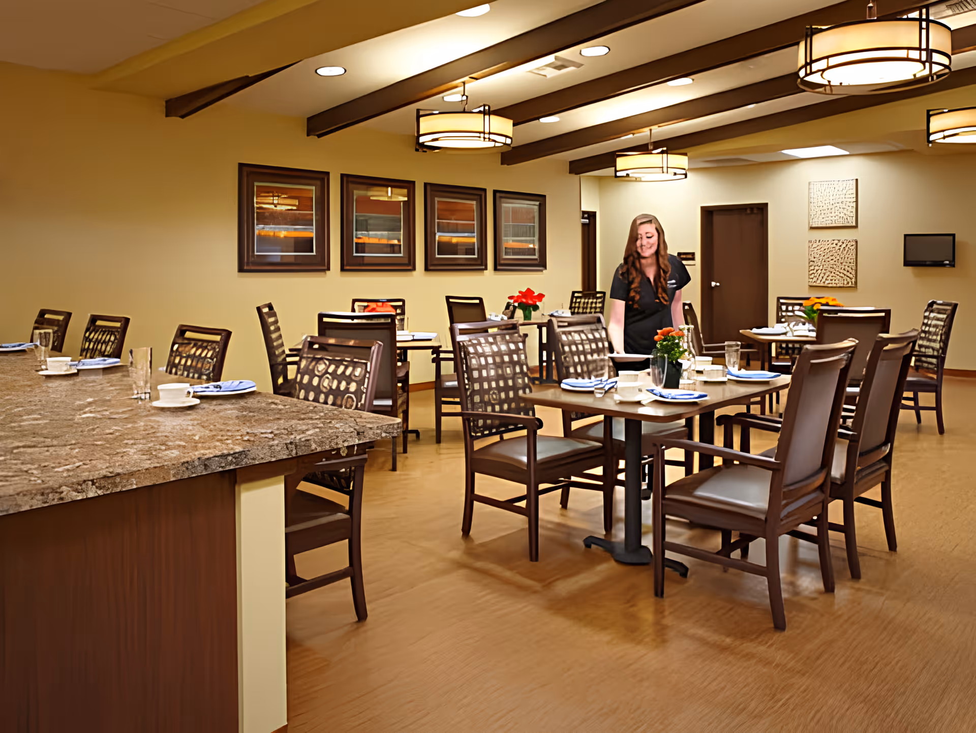 Well-lit dining room with multiple set tables and chairs, a staff member arranging a table, and a marble countertop in the foreground.