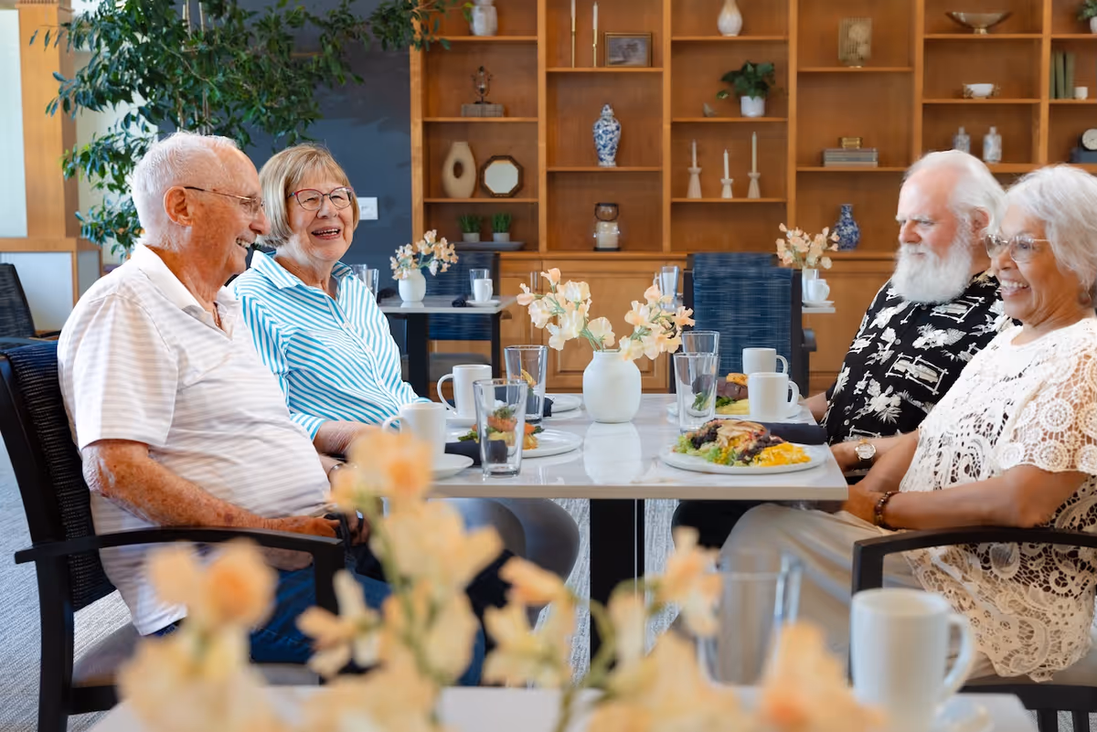Four senior residents sit and laugh around a dining table with plates and flower centerpieces in a communal dining room.